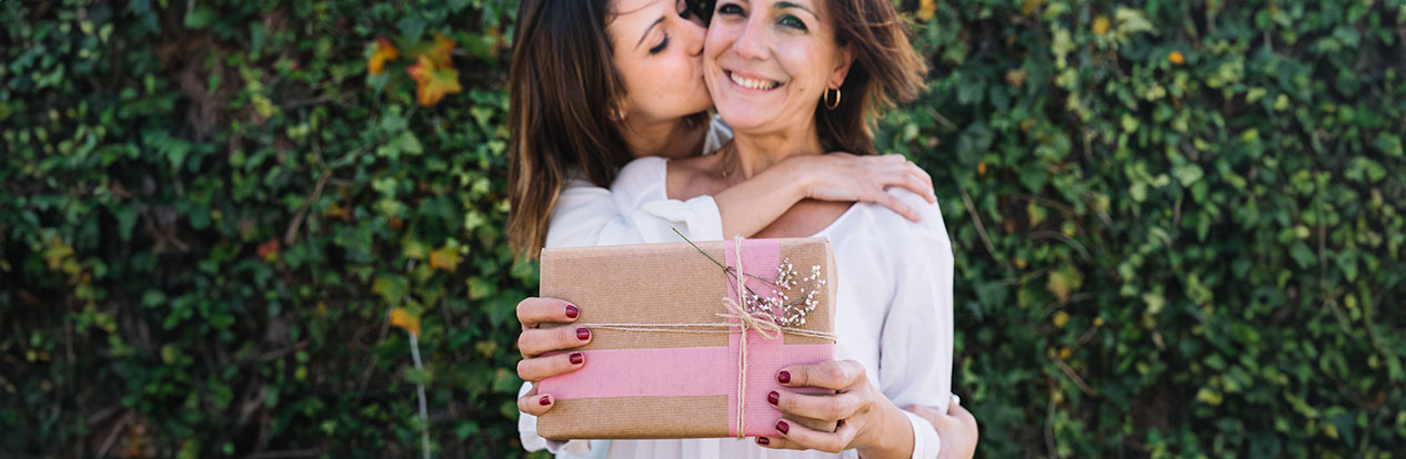 Jovem mulher beijando a bochecha da sua mãe, que sorri felizmente, enquanto segura um presente embrulhado com papel kraft e decorado com uma fita rosa e um ramo pequeno, em um fundo de folhas verdes.
